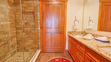 A bathroom with a wooden door, a glass shower enclosure, a granite countertop, and a red patterned rug.