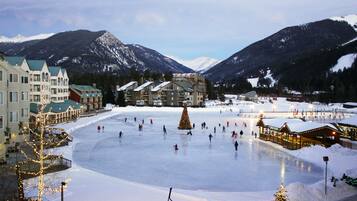 A large outdoor ice rink surrounded by buildings with balconies, a Christmas tree, and snow-covered mountains in the background.