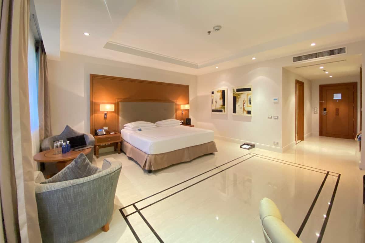 A modern hotel room with a large bed, a seating area with a chair, a desk with a laptop, and artwork on the walls.