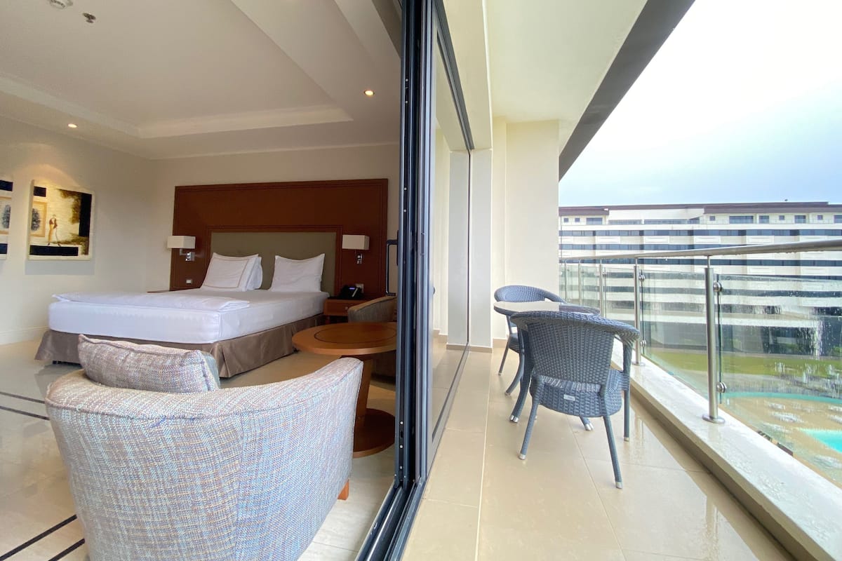 A hotel room with a bed, a small table, two chairs, and a balcony with a view of buildings and a pool.