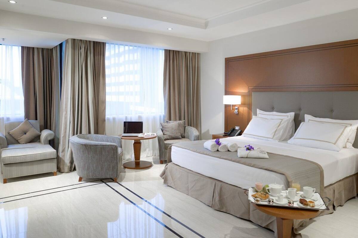 A hotel room with a large bed, a desk, a chair, and a dining table with breakfast items.