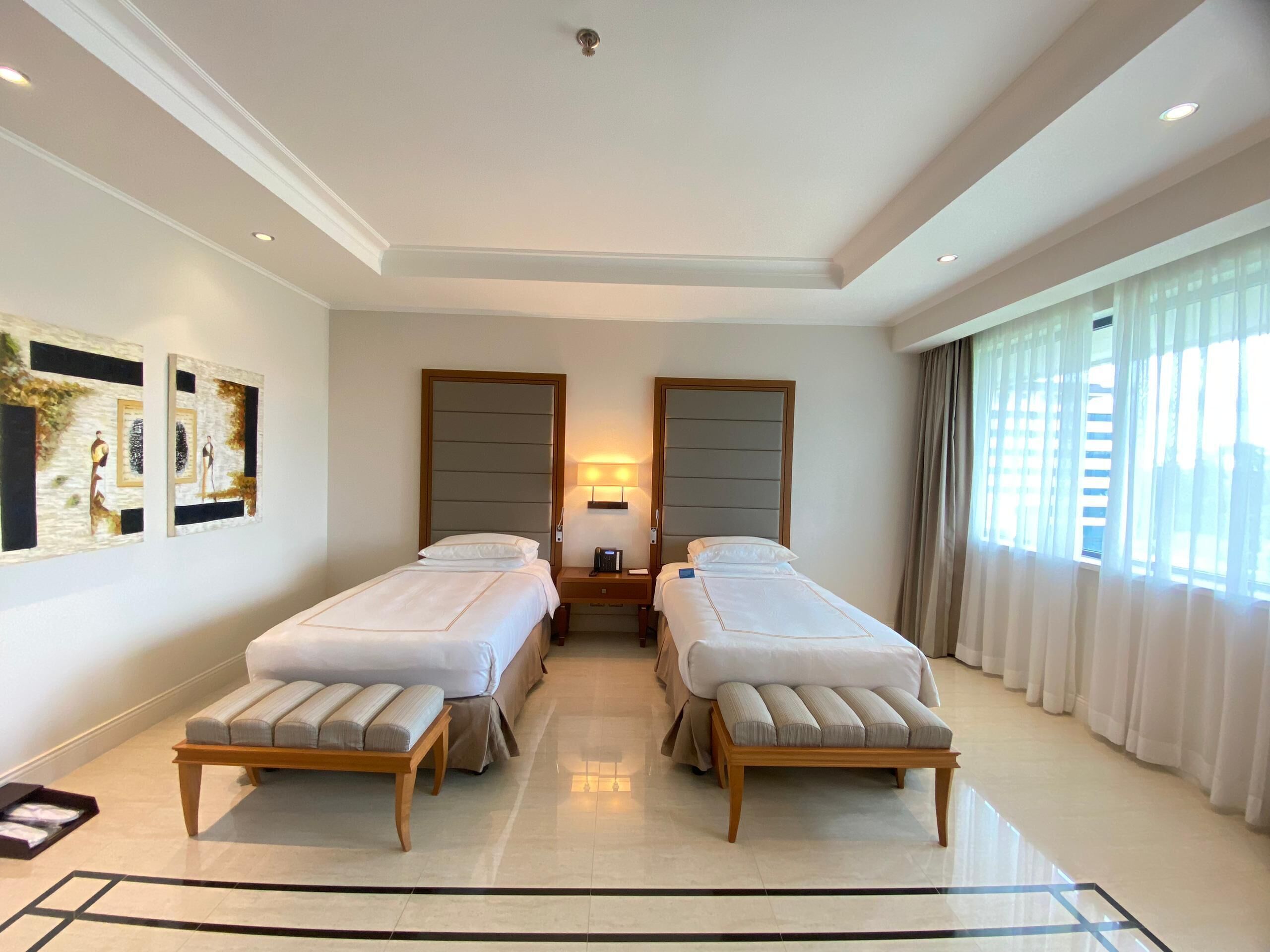 A hotel room with two single beds, a bedside table with a lamp, a large window with curtains, and artwork on the wall.