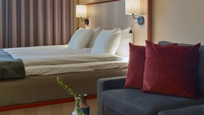 A hotel room with a neatly made bed, a grey sofa, and a small table with books and a vase.