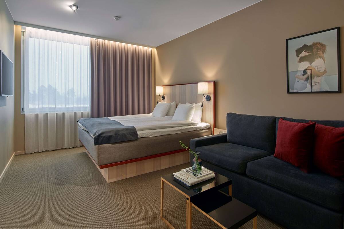 Una habitación de hotel con cama, sofá, una mesita y un ventanal con cortinas.