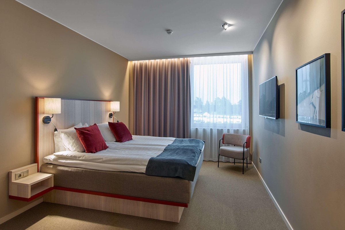 Una habitación de hotel con una cama grande, lámparas de noche, una silla, televisión y ventana con cortinas.