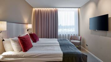 Ein Hotelzimmer mit einem großen Bett, zwei Kissen, einem Sessel, einem Fernseher und einem Fenster mit Vorhängen.