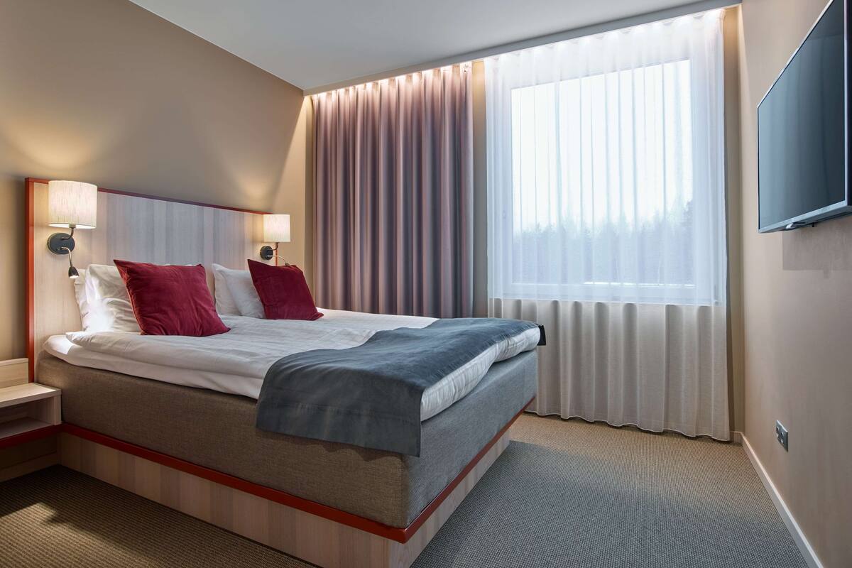 Habitación de hotel con cama, lámparas de noche, televisión y ventana con cortinas.