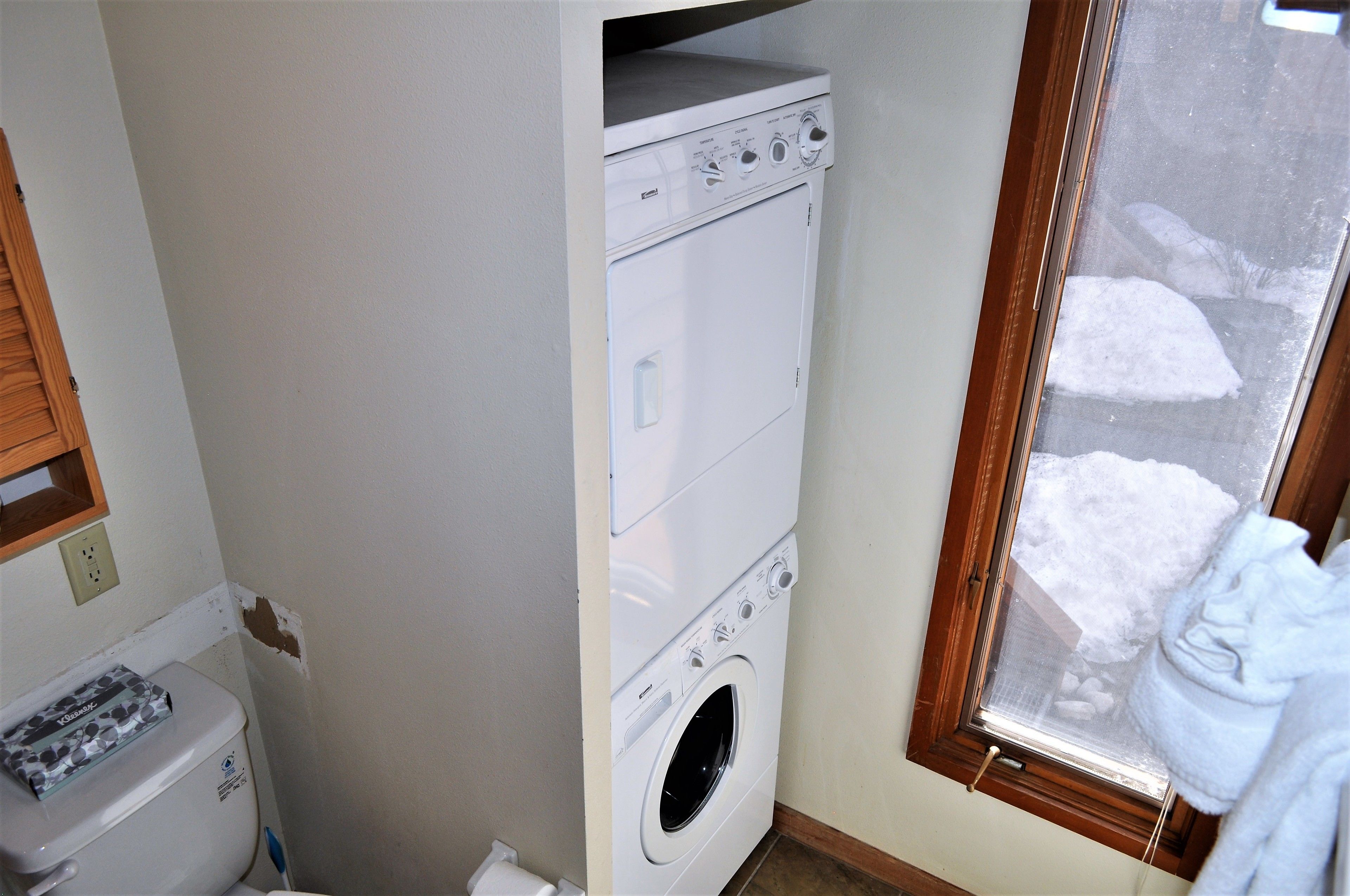 A laundry room with a stacked washer and dryer, a window with a view of snow outside, and a toilet with a paper towel dispenser.