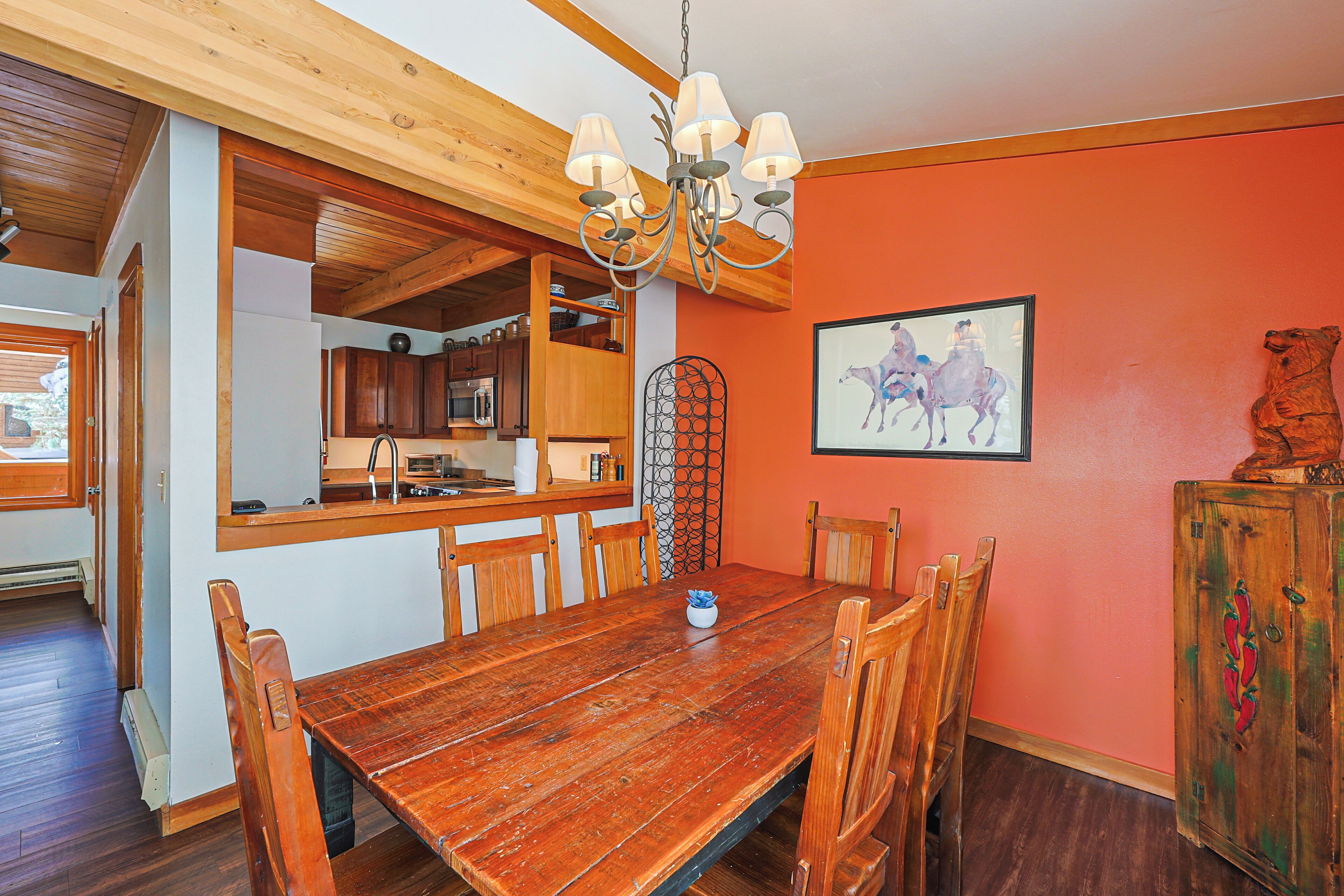 A dining area with a wooden table and chairs, a chandelier, and a kitchen in the background.