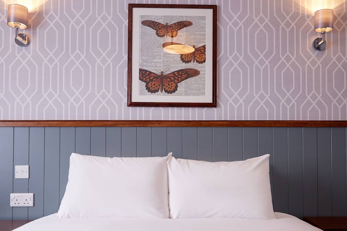 Een net opgemaakt bed met witte lakens en een grijze deken, een lijst met een vlinderillustratie aan de muur, en twee wandlampen.