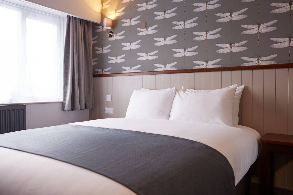 Een hotelkamer met een groot bed, een raam met gordijnen, een muur met een libellenprint en een houten hoofdeinde.