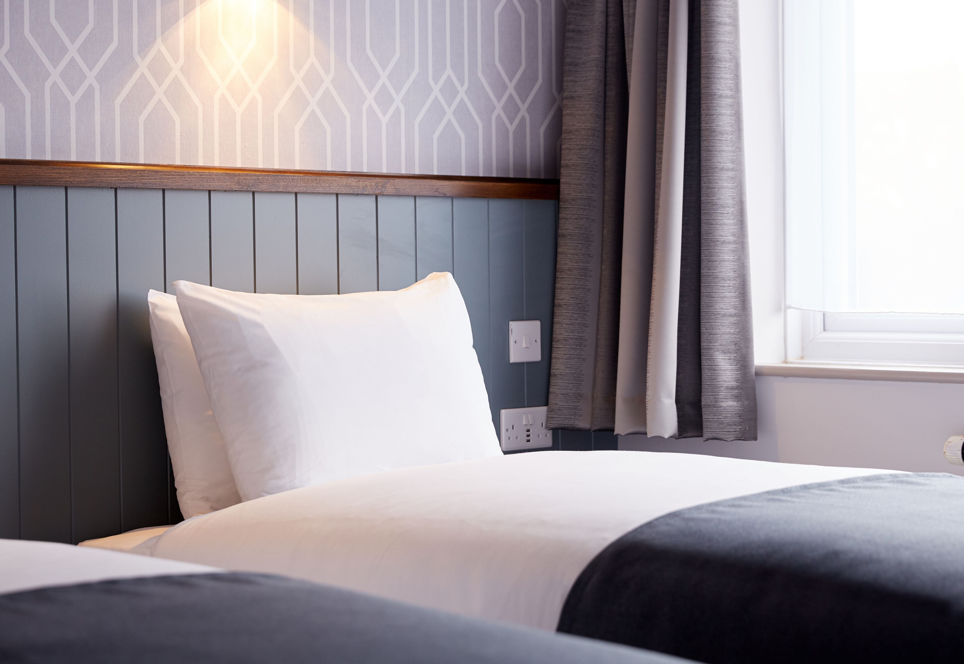 A hotel room with a bed, a headboard with a wooden panel, a wall with a geometric pattern, and a window with curtains.