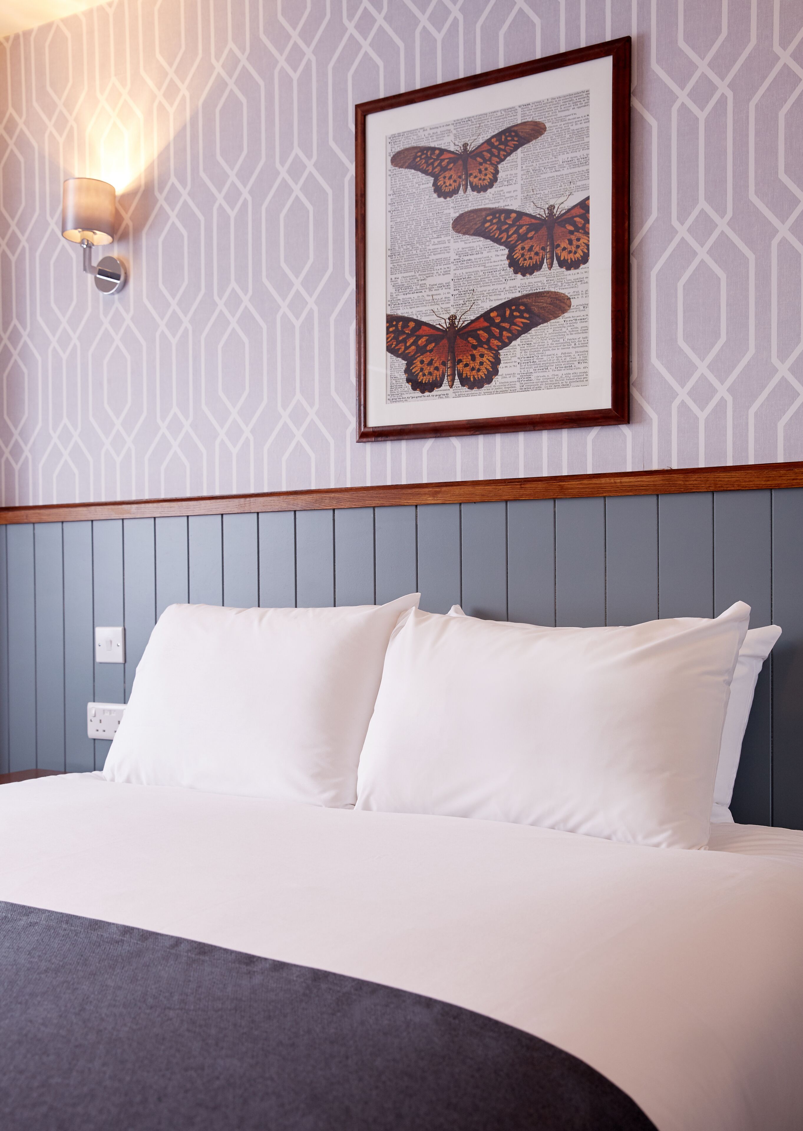 A neatly made bed with white linens and a grey headboard. Above the bed, there is a framed picture of butterflies on a patterned wall.