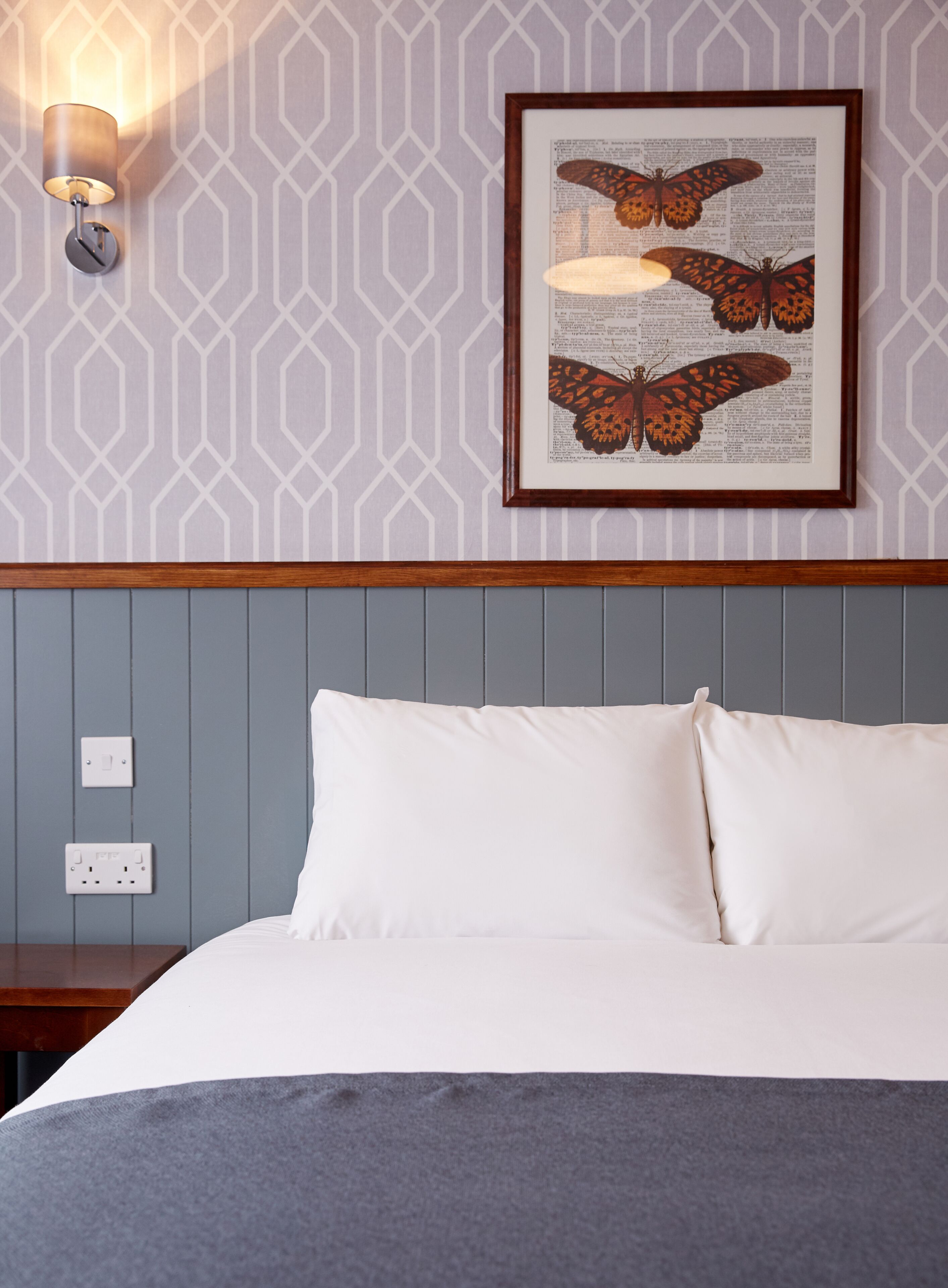 A neatly made bed with white linens, a framed butterfly artwork on the wall, and a wall-mounted light fixture.