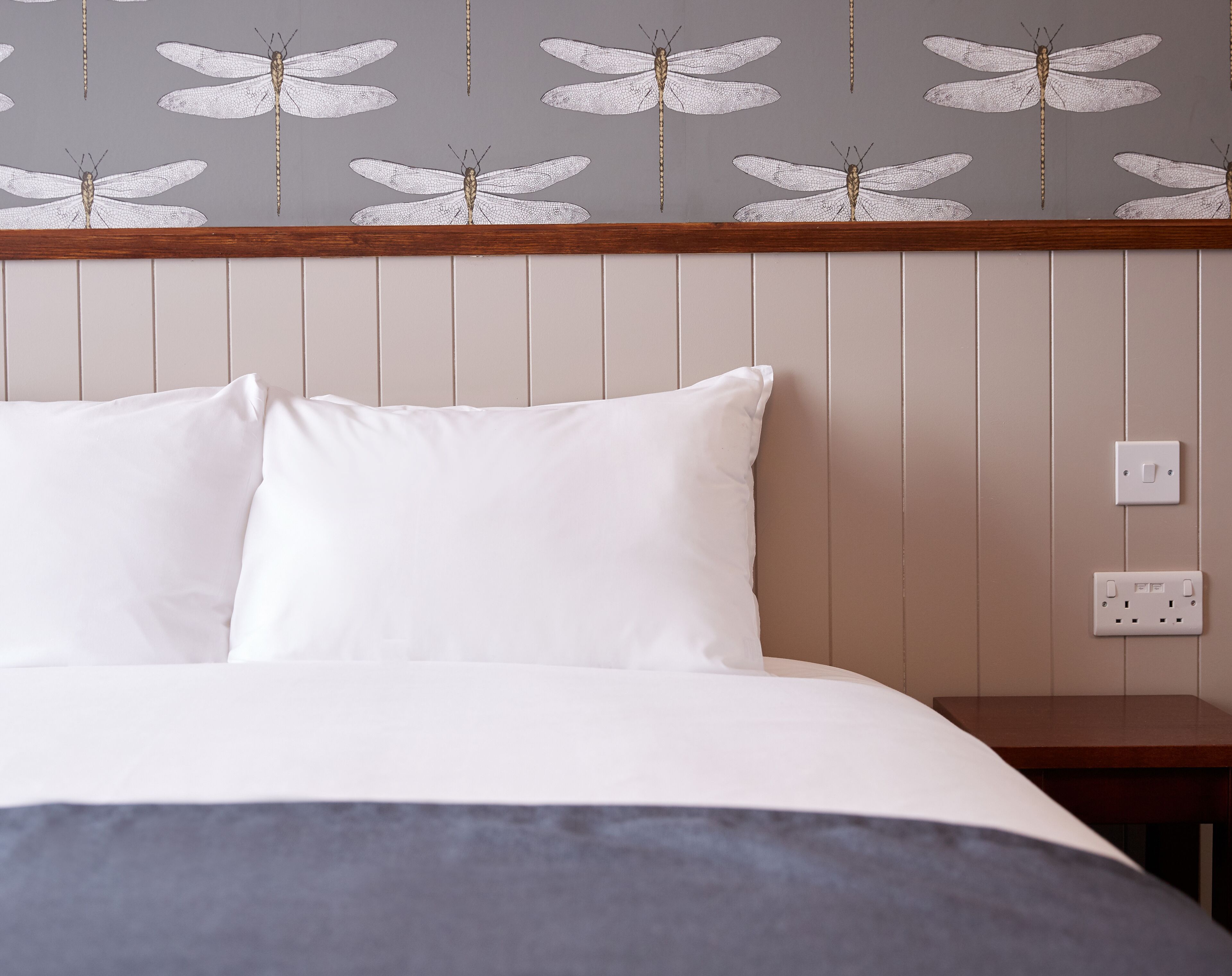 A bed with white bedding and a grey blanket, a wooden headboard, and a wall with a dragonfly pattern.