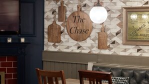 A dining area with a wooden table and chairs, a bench, and a wall with a geometric tile pattern and a sign that reads "The Chase". - The Chase Hotel by Greene King Inns (Nuneaton)
