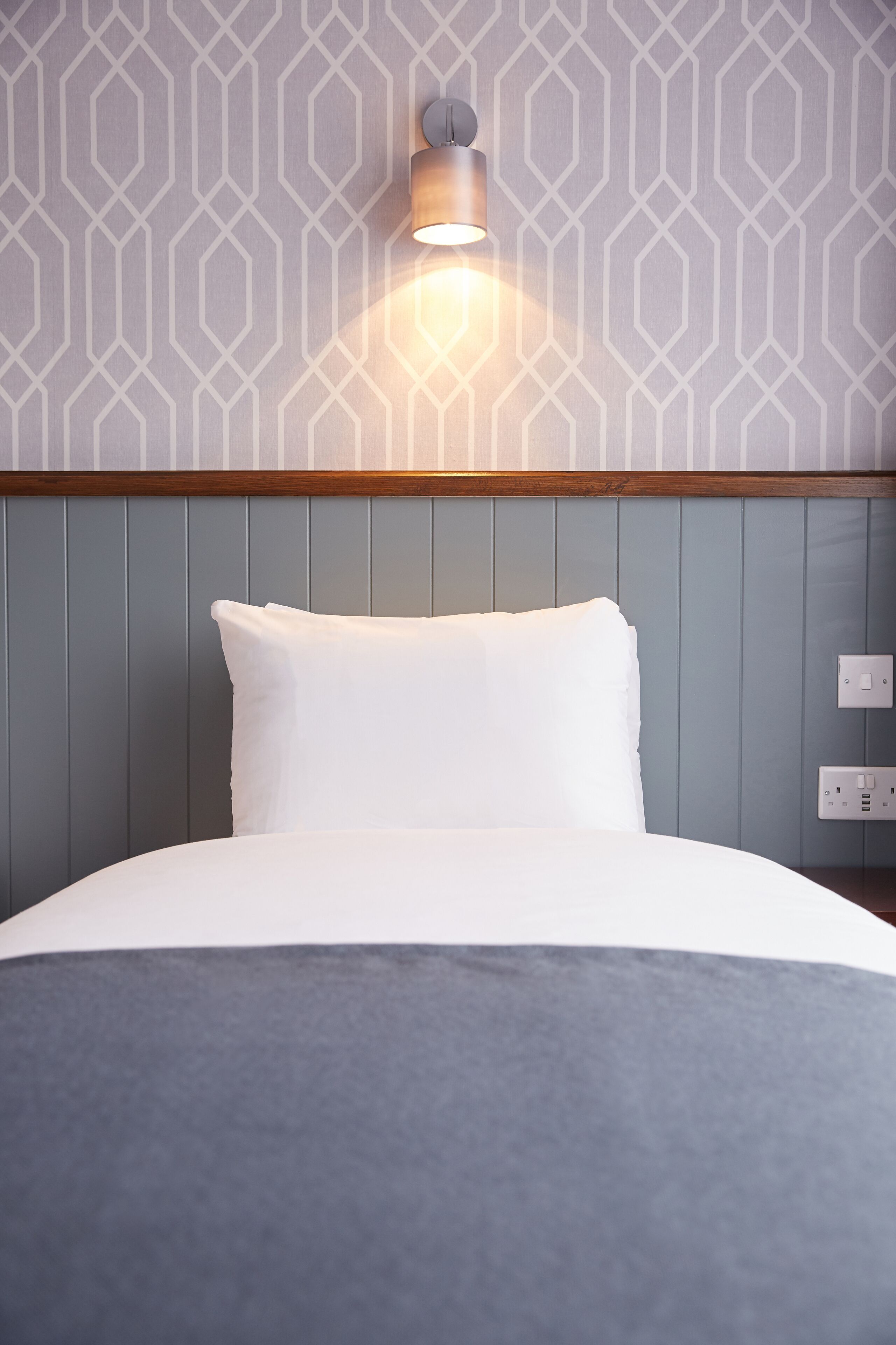 Cama bem arrumada com travesseiro branco e manta cinza, luminária fixada na parede e tomada elétrica em parede com padrão geométrico.