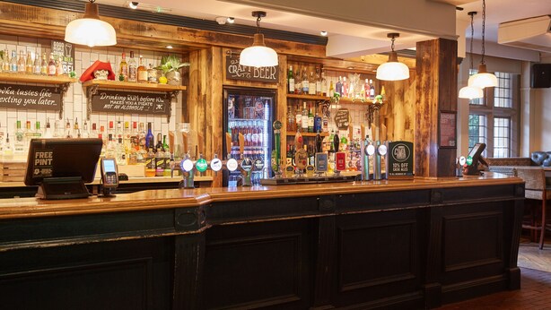 A bar area with a wooden counter, multiple beer taps, and a variety of liquor bottles. - The Chase Hotel by Greene King Inns (Nuneaton)