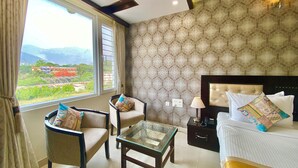 A hotel room with a large window, a bed, a chair, a small table, and a nightstand with a lamp. - Hotel Green View - Behind Pramarth Niketan (Yamkeshwar)