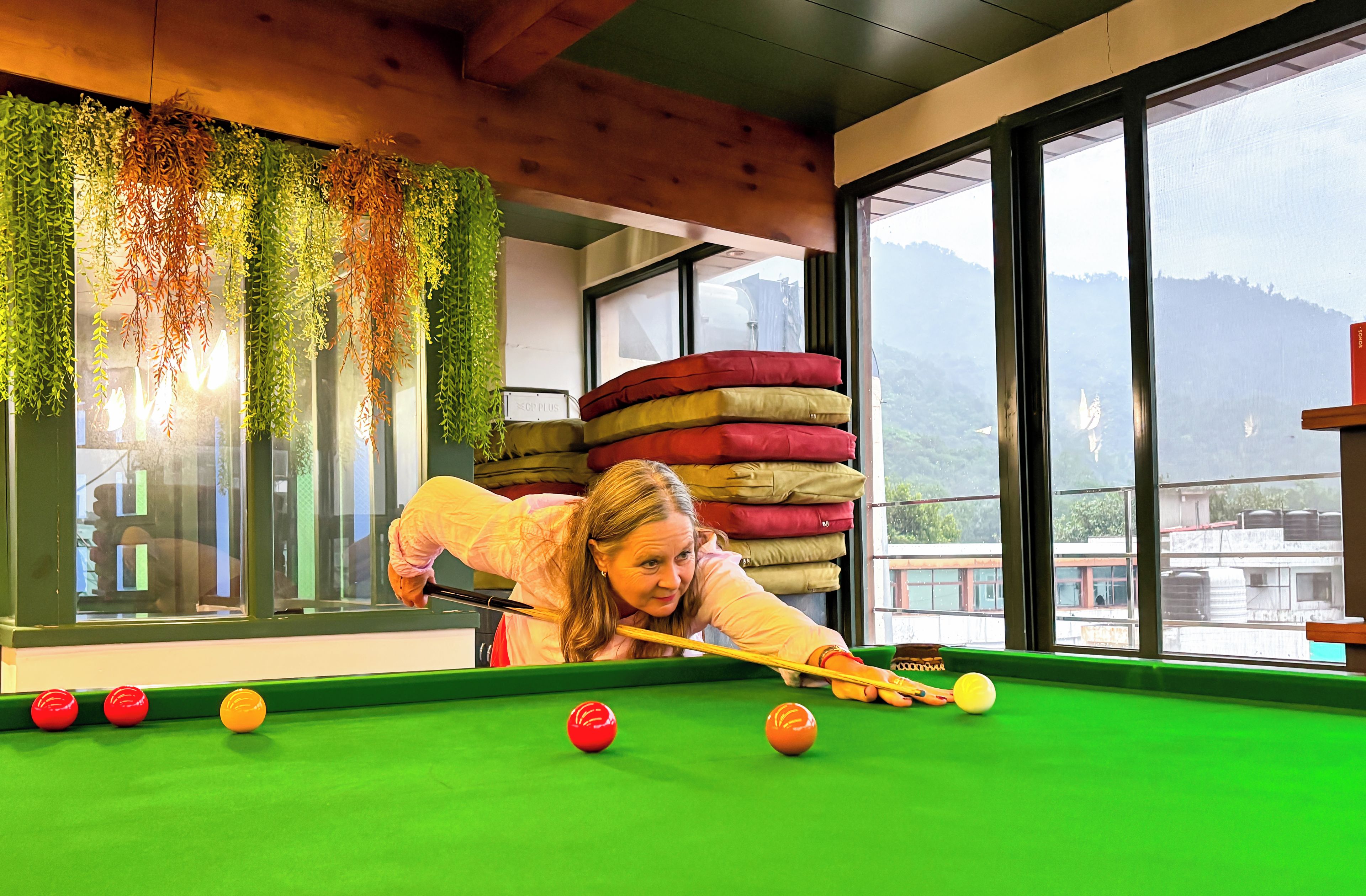 Una mesa de billar con bolas, una persona jugando y una ventana con vistas a edificios y montañas.