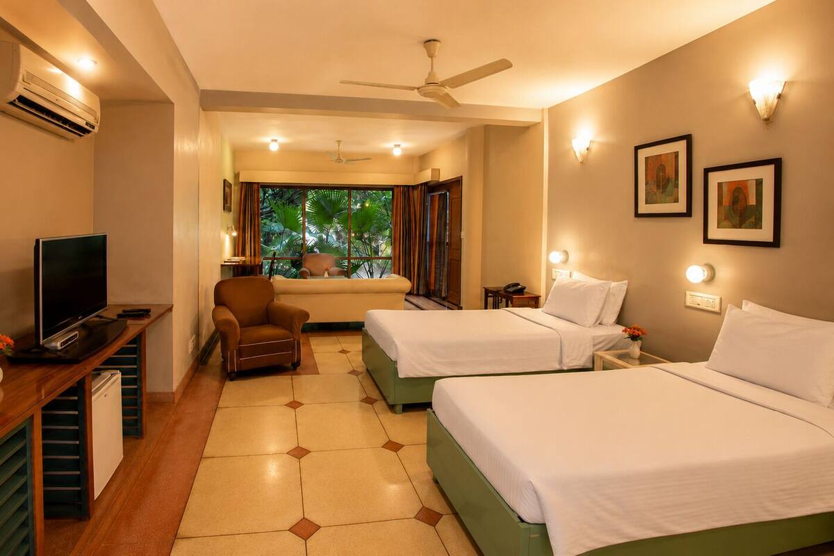 A hotel room with a TV, two beds, a chair, a desk, and a view of greenery outside.