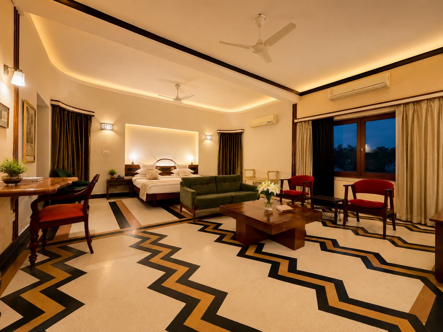 A hotel room with a bed, green sofa, red chairs, a coffee table, and a view of the outdoors.
