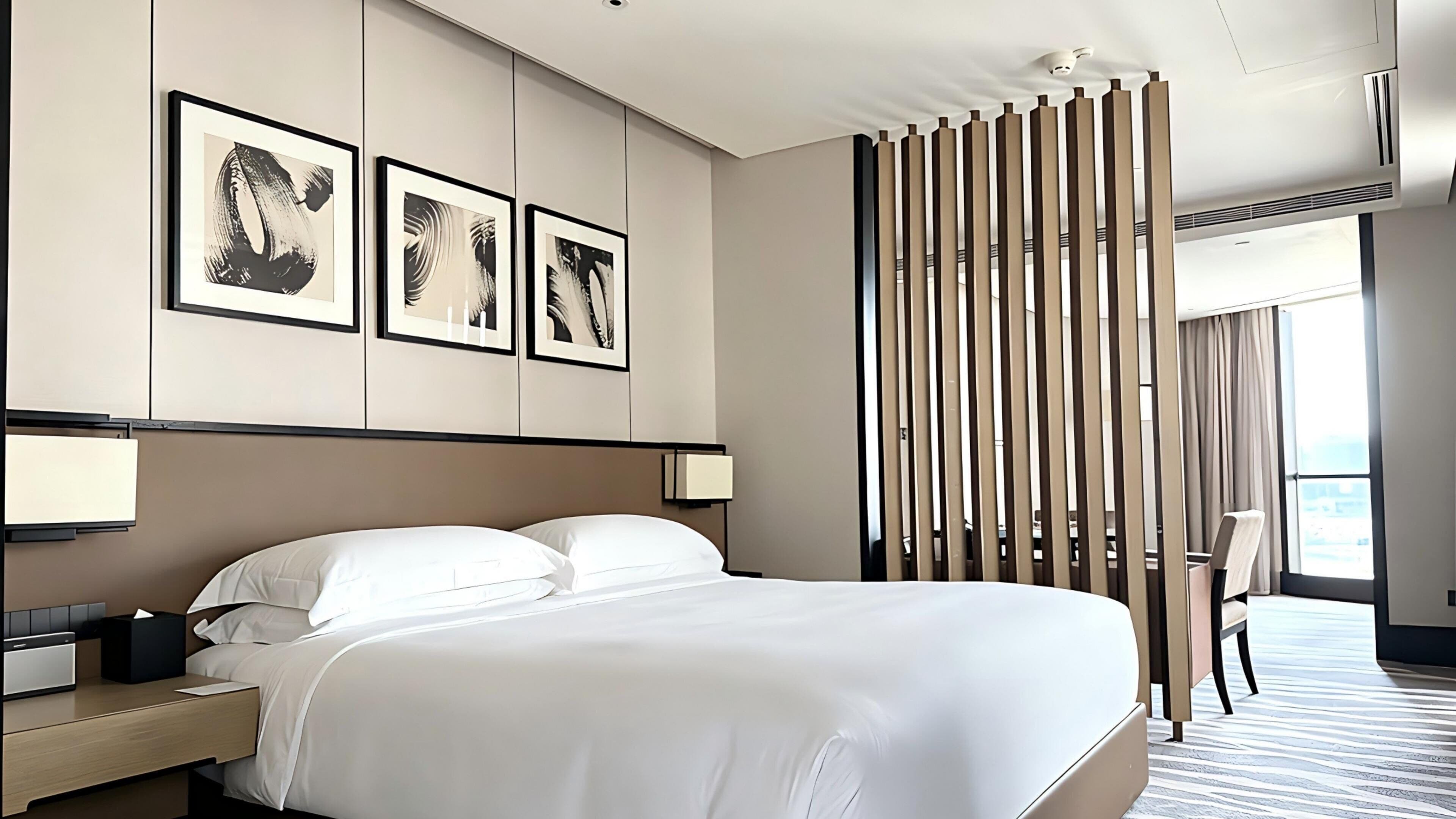 Ein Hotelzimmer mit einem großen Bett, Nachttischen, einem Schreibtisch und einem Sessel. An der Wand hängen drei gerahmte Kunstwerke.