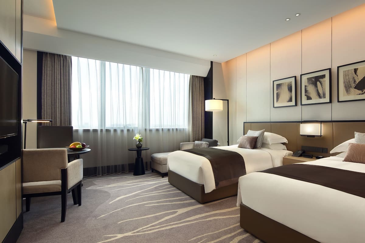 A hotel room with two beds, a desk with a fruit bowl, a chair, and a TV.