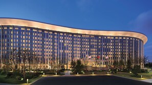 A modern building with a curved facade, multiple windows, and a well-lit entrance area with trees and a pathway. - InterContinental Shanghai Hongqiao NECC by IHG (Shanghai)