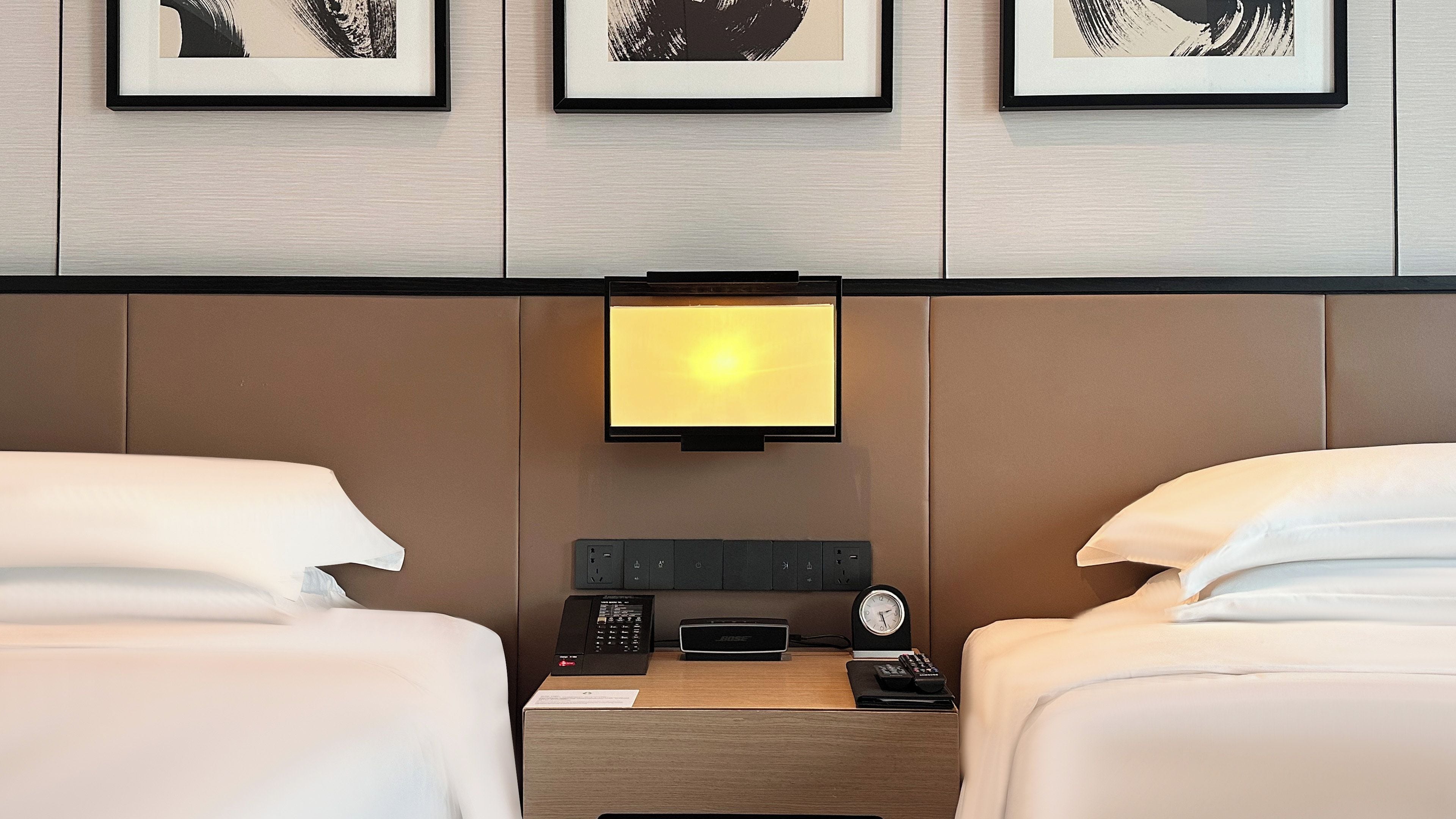 A hotel room with two beds, a bedside table with a clock, a remote control, and a speaker.