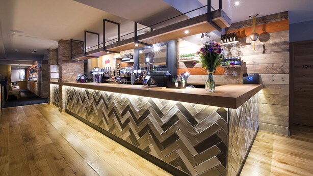 Een bargedeelte met een houten toonbank, barstoelen en diverse flessen en glazen.