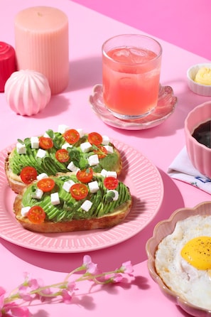 Ett rosa bord med en tallrik avokadotoast, ett glas rosa dryck och en skål smör.