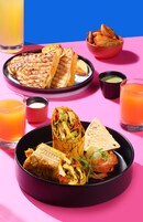 Una mesa con diversos alimentos, como sándwiches a la parrilla, un wrap y un bol de papas fritas, junto con vasos de jugo de naranja.
