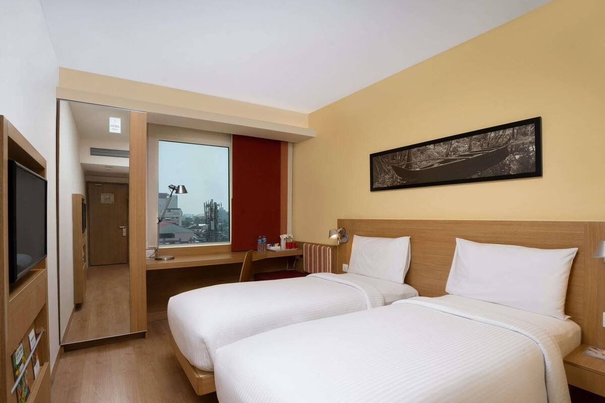 A hotel room with two beds, a TV, a desk, and a window with a city view.