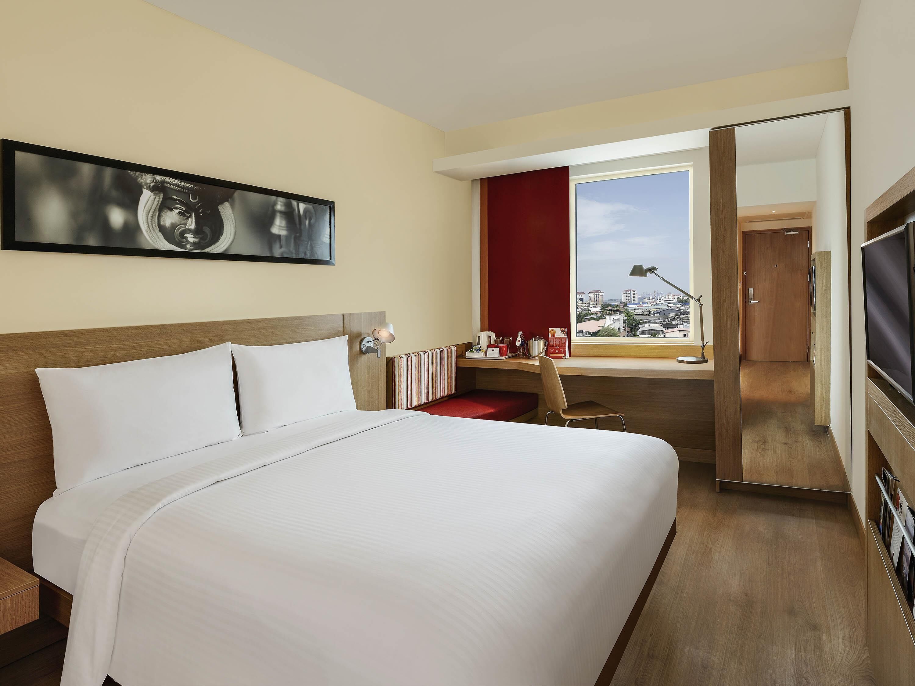 Ein Hotelzimmer mit einem großen Bett, einem Schreibtisch mit Stuhl, einem Fenster mit Blick auf die Stadt und einem Fernseher.