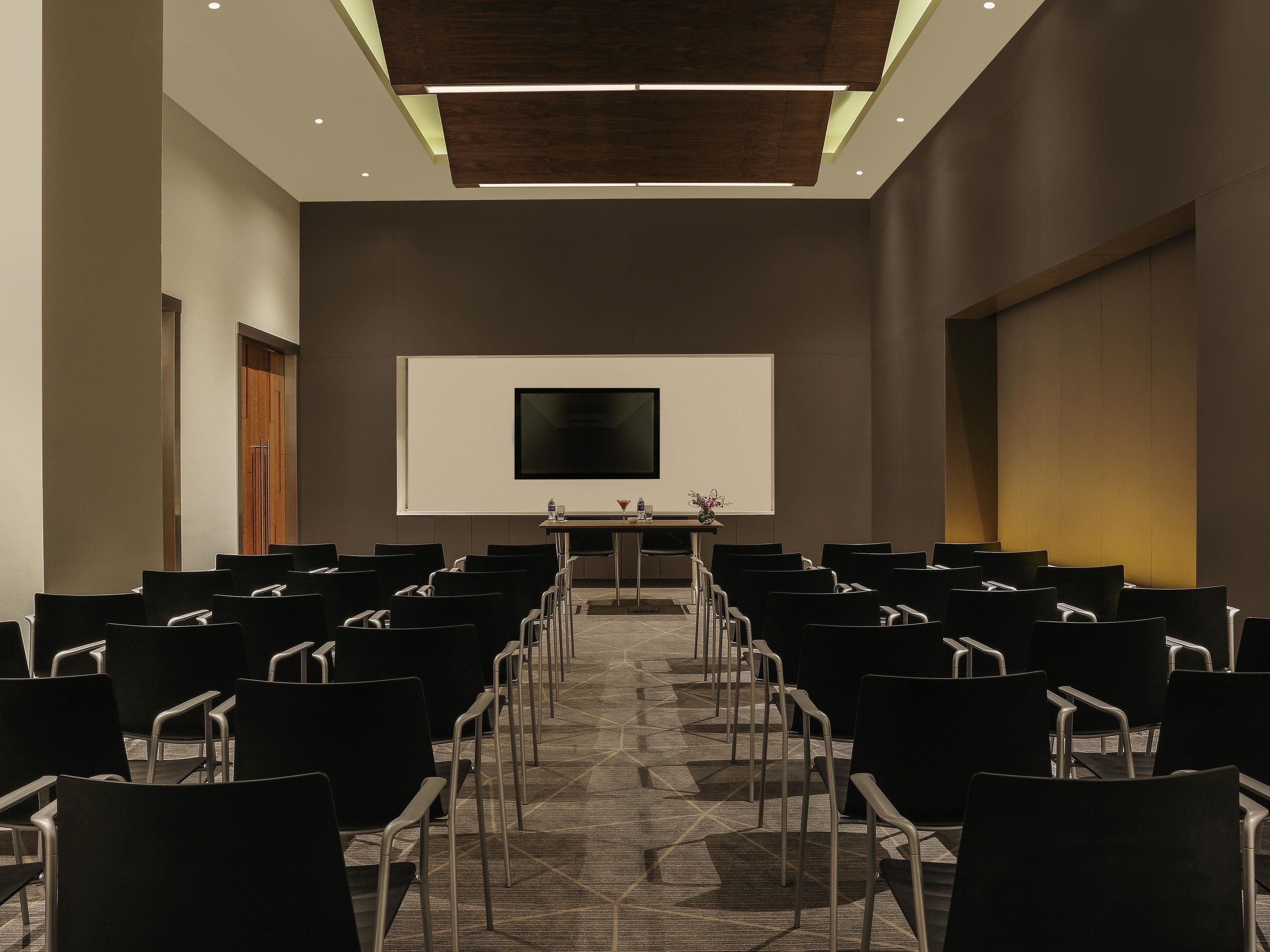 Un salón de conferencias con filas de sillas, una pantalla de proyector y una mesa con una botella de agua y una flor.