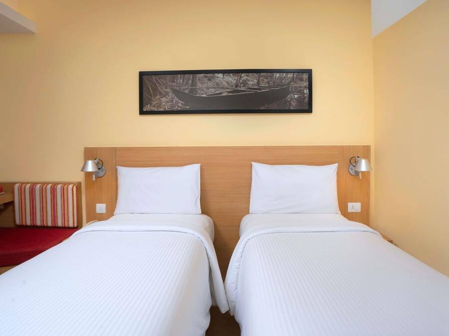 Habitación de hotel con dos camas, cabecero de madera, un cuadro colgado en la pared y un banco rojo.