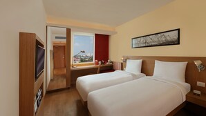 Ein Hotelzimmer mit zwei Betten, einem Fernseher, einem Schreibtisch mit Stuhl, einem Fenster mit Blick auf die Stadt und einem gerahmten Bild an der Wand.