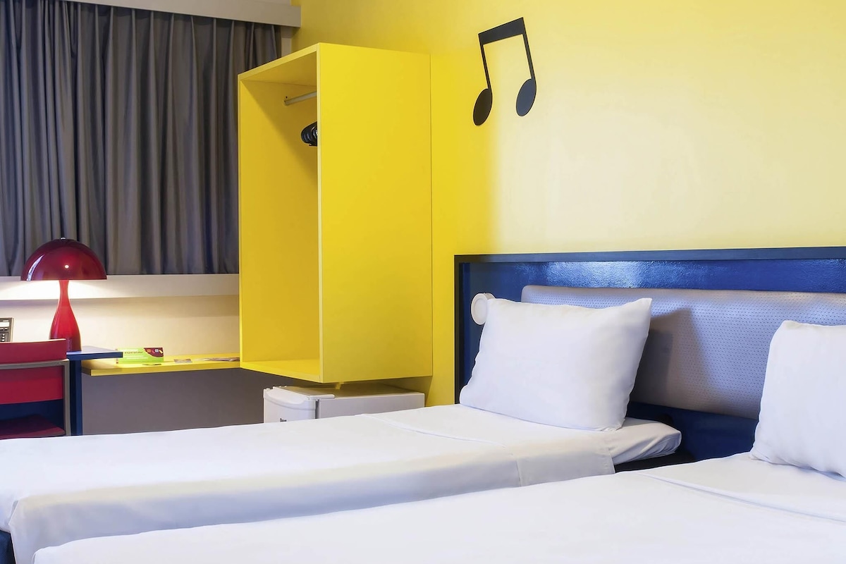 Una habitación de hotel con dos camas, un escritorio, una lámpara roja y un armario amarillo.