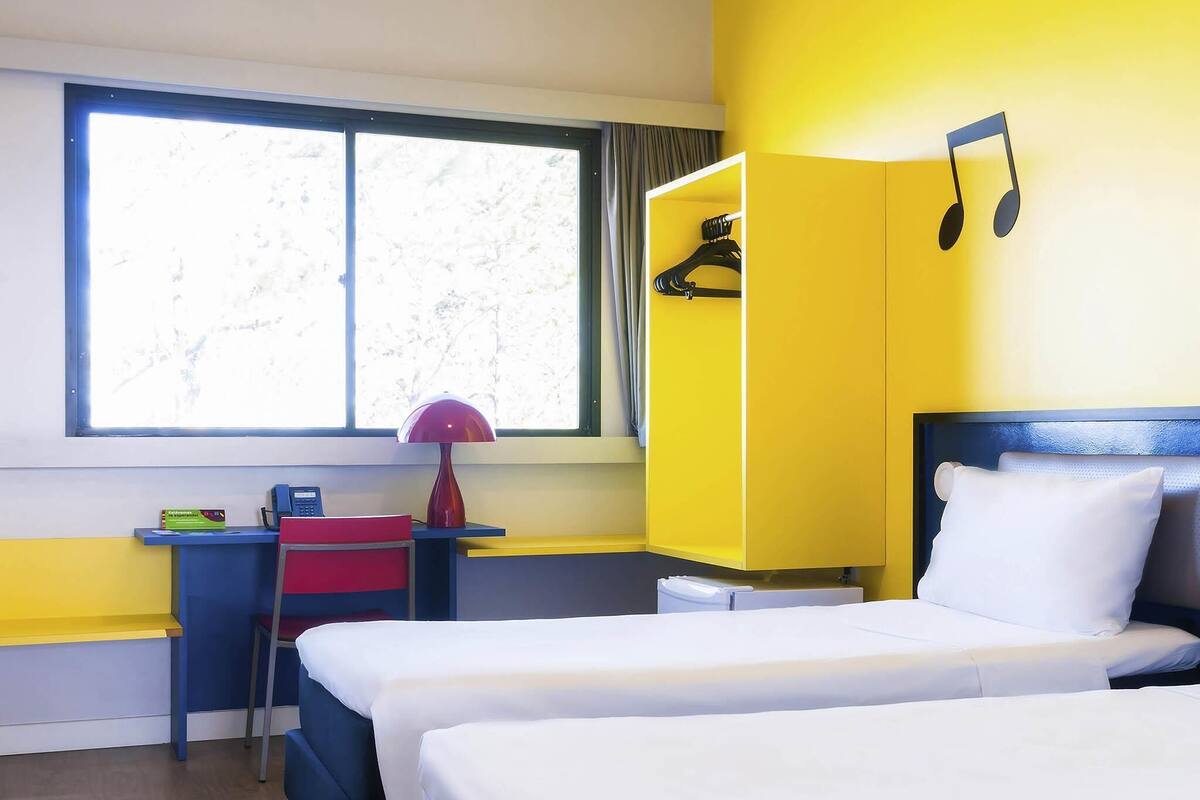 Quarto de hotel com duas camas, uma escrivaninha com cadeira, uma luminária e um guarda-roupa.