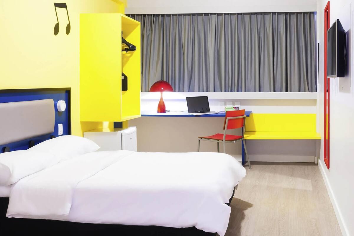 Quarto de hotel com uma cama, uma escrivaninha com um laptop, uma cadeira e uma televisão na parede.