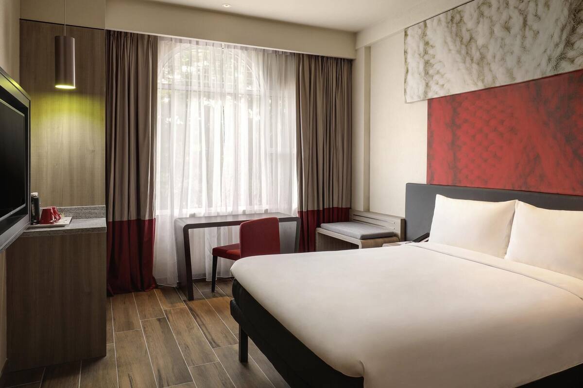 A hotel room with a bed, a desk, a chair, a TV, and a window with curtains.