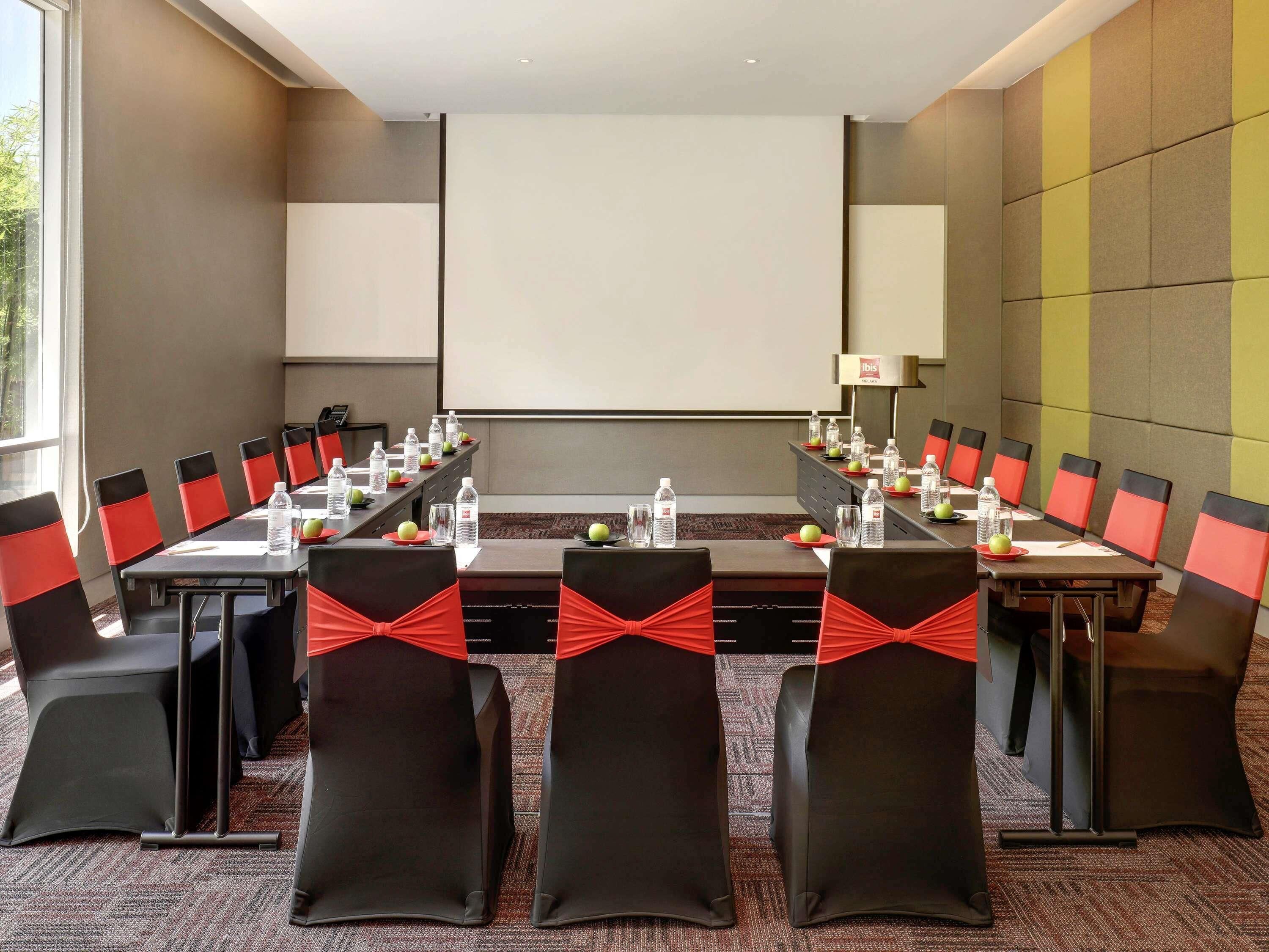 Un salón de conferencias con una larga mesa, sillas con cintas rojas, botellas de agua y manzanas sobre la mesa.