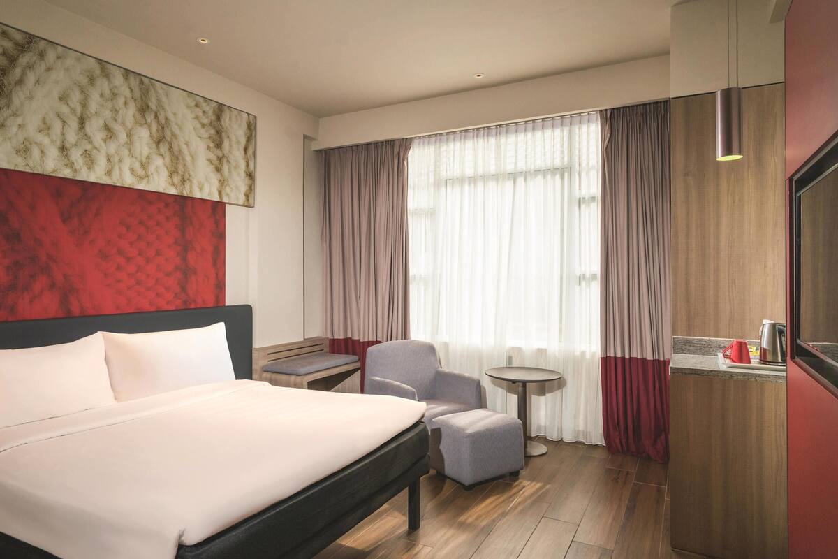 A hotel room with a large bed, a small table, a chair, and a desk with a kettle.