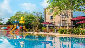 A swimming pool with a water slide and play structures, surrounded by trees and a building with outdoor seating. - ibis Melaka (Malacca City)