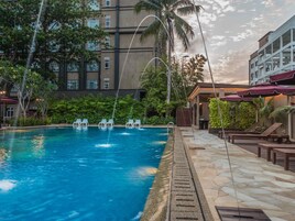 Una piscina con chorros de agua, rodeada de sillones de descanso y un edificio de varios pisos.