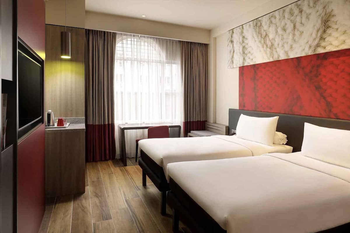 A hotel room with two beds, a desk, a television, and a large window with curtains.