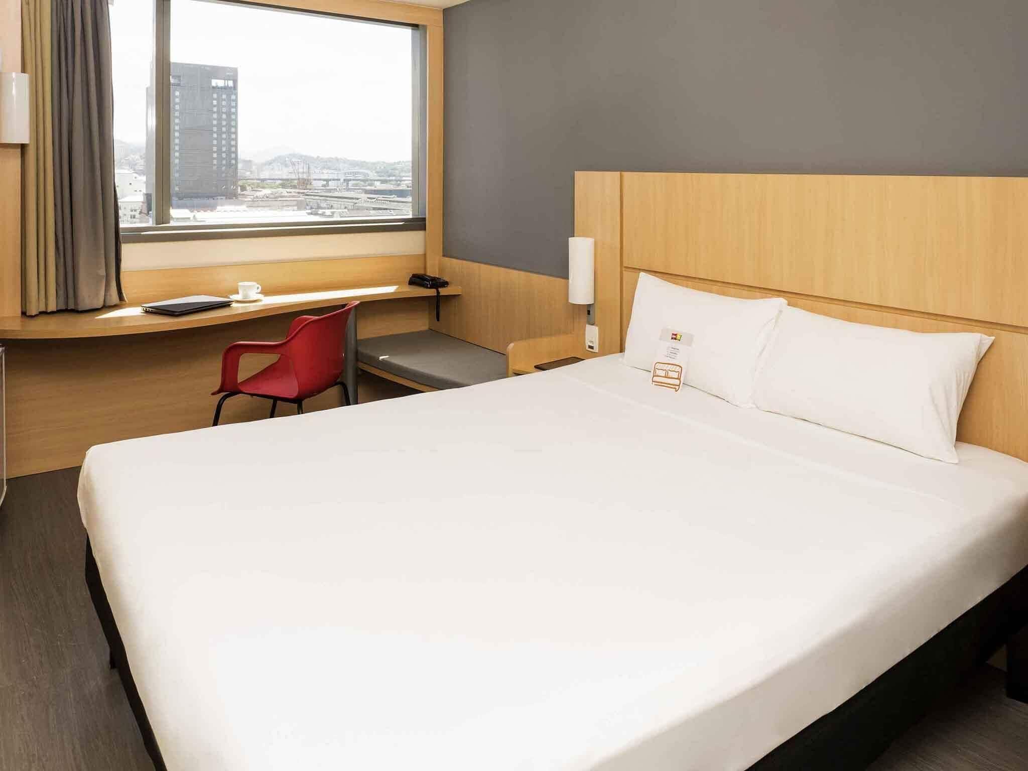 A hotel room with a large bed, a desk, a red chair, a window with a city view, and a wooden headboard.