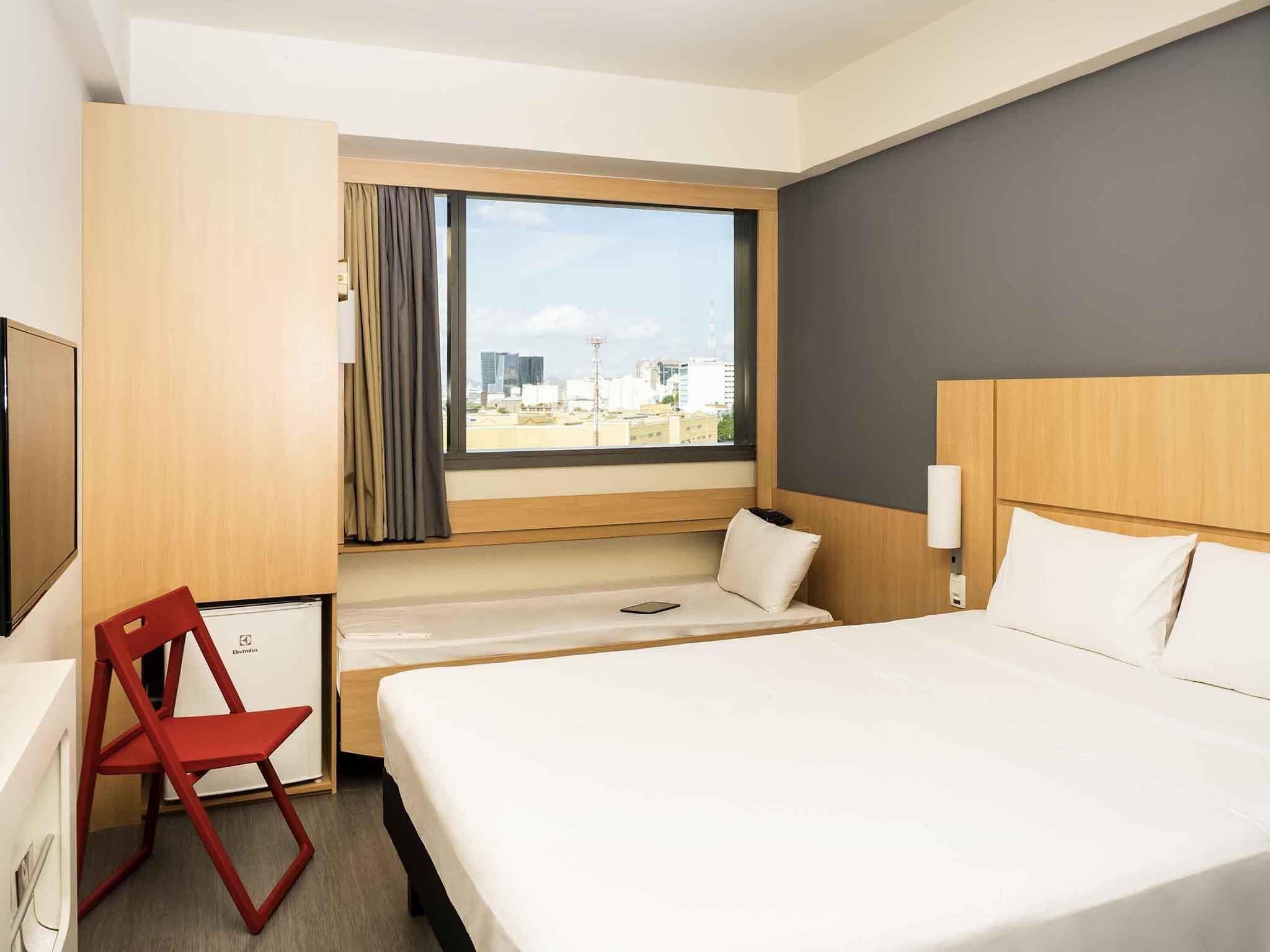 A hotel room with a bed, a desk, a chair, a window with a city view, and a wall-mounted lamp.
