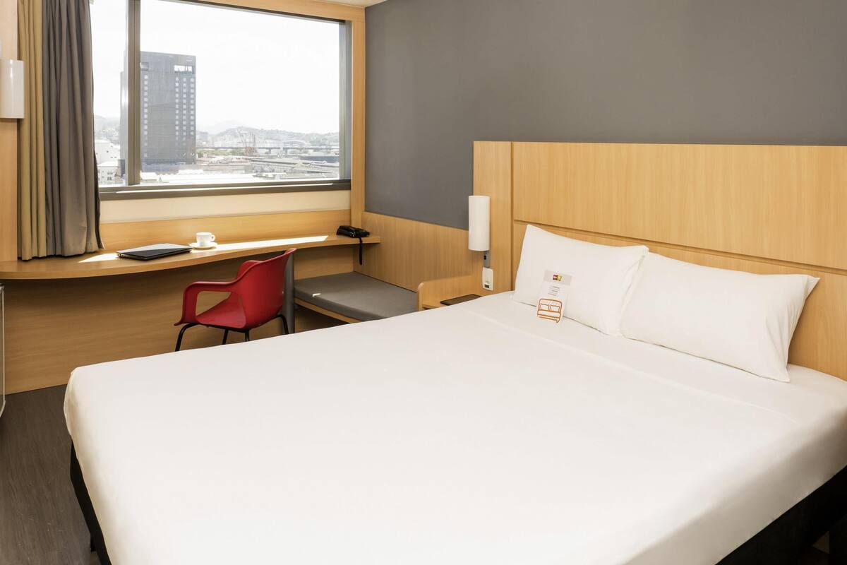 A hotel room with a large bed, a desk, a red chair, a window with a view of buildings, and a bedside table with a lamp.